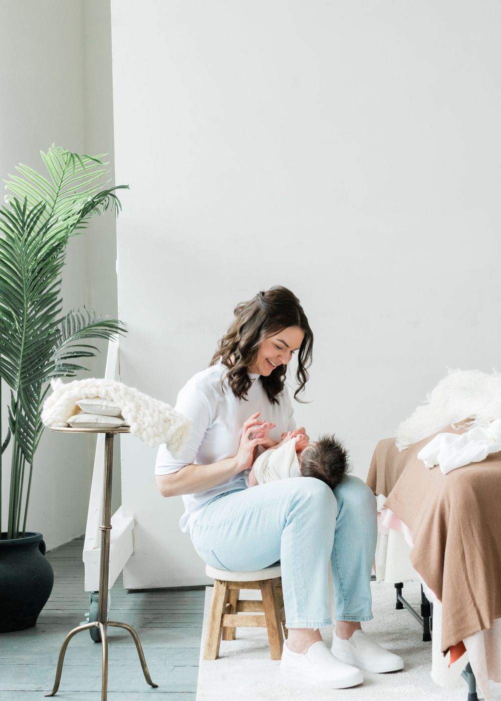 A person in a white sweater and light blue pants sitting on a wooden stool next to a potted palm plant.