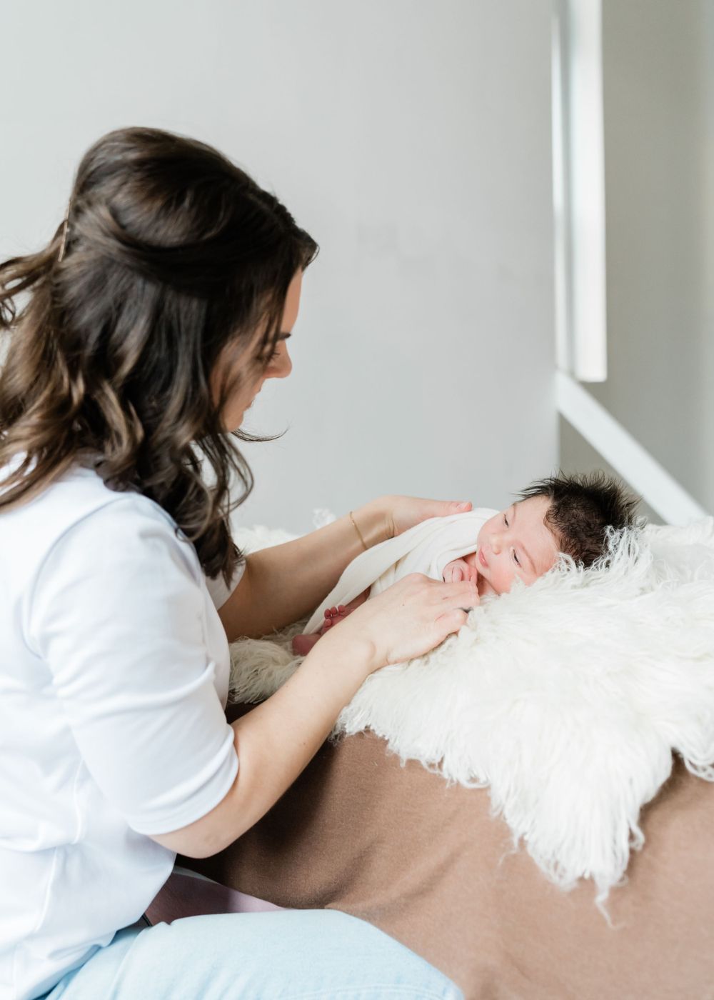 A woman in white gently cares for a newborn baby resting on plush white bedding.