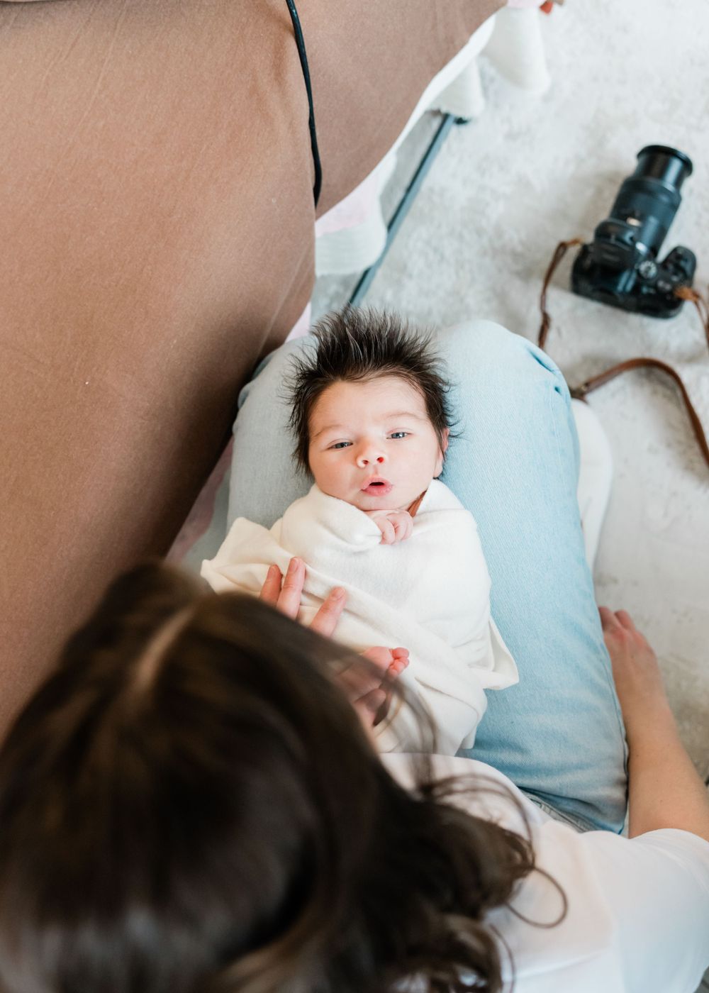 A baby with spiky dark hair lies on a light blue pillow while being photographed from above.