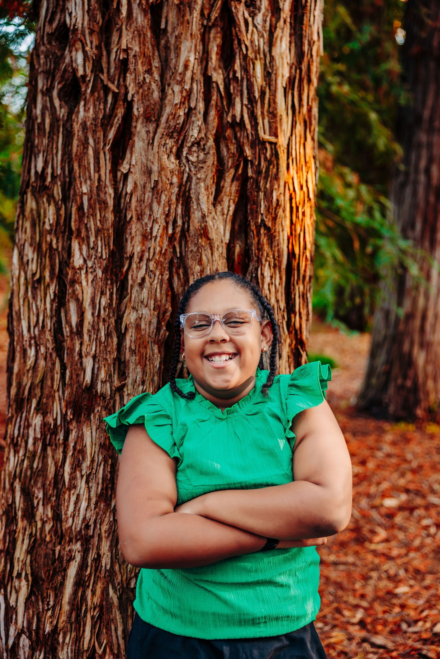 Person in vibrant green shirt poses against redwood tree trunk with fall leaves scattered on ground.