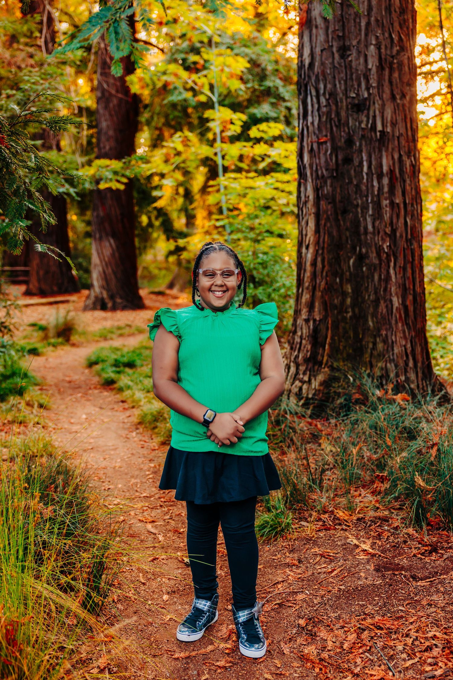 Standing on a forest trail wearing a bright green top and navy skirt among tall redwood trees in autumn.