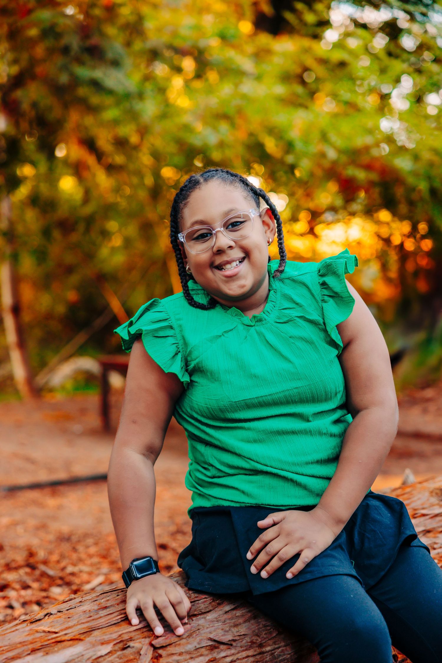A person in a green shirt and dark pants sits among autumn leaves on the ground while smiling warmly.