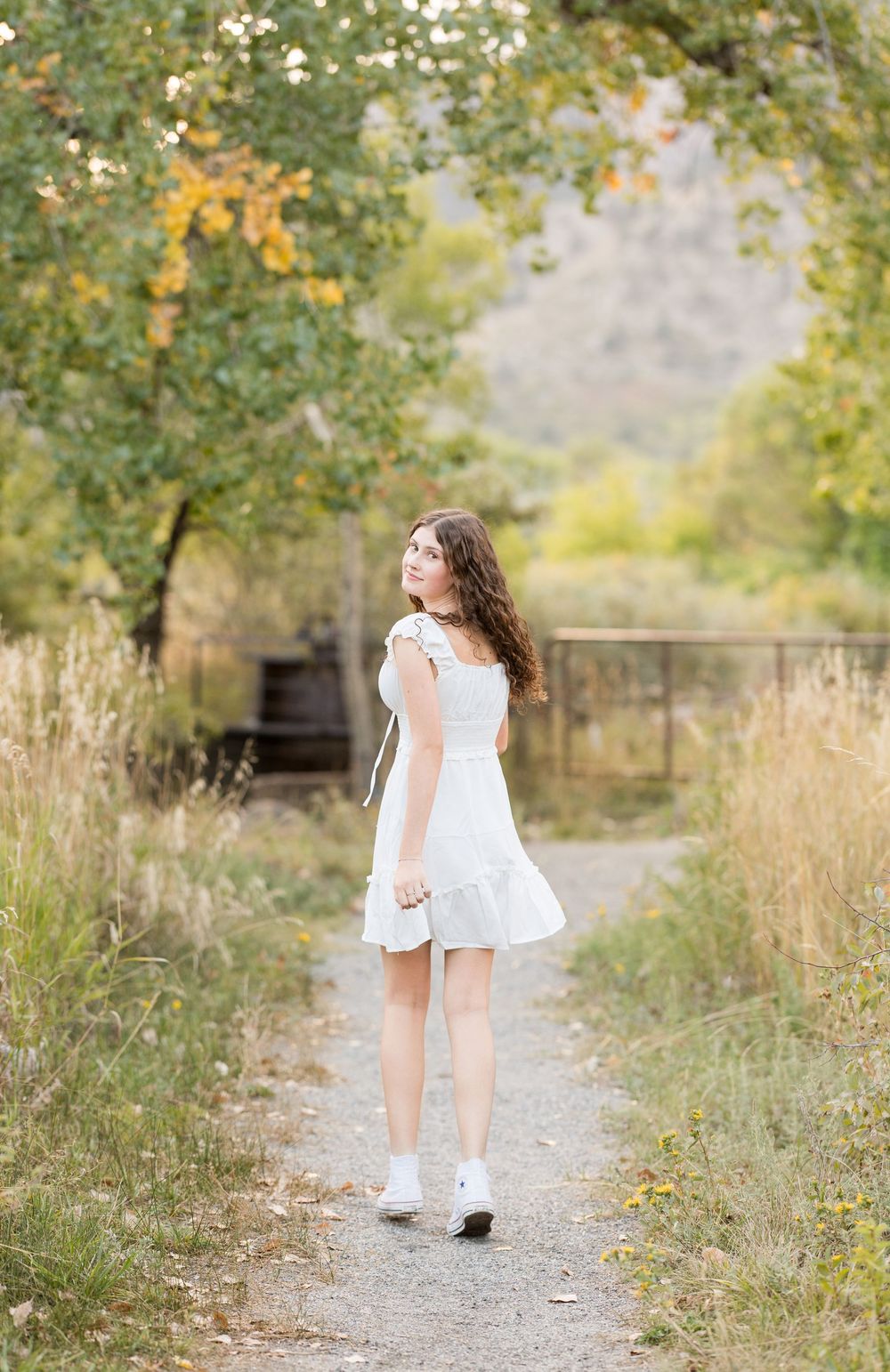 A white dress flows gracefully along a sunlit nature path bordered by tall grass and autumn trees.