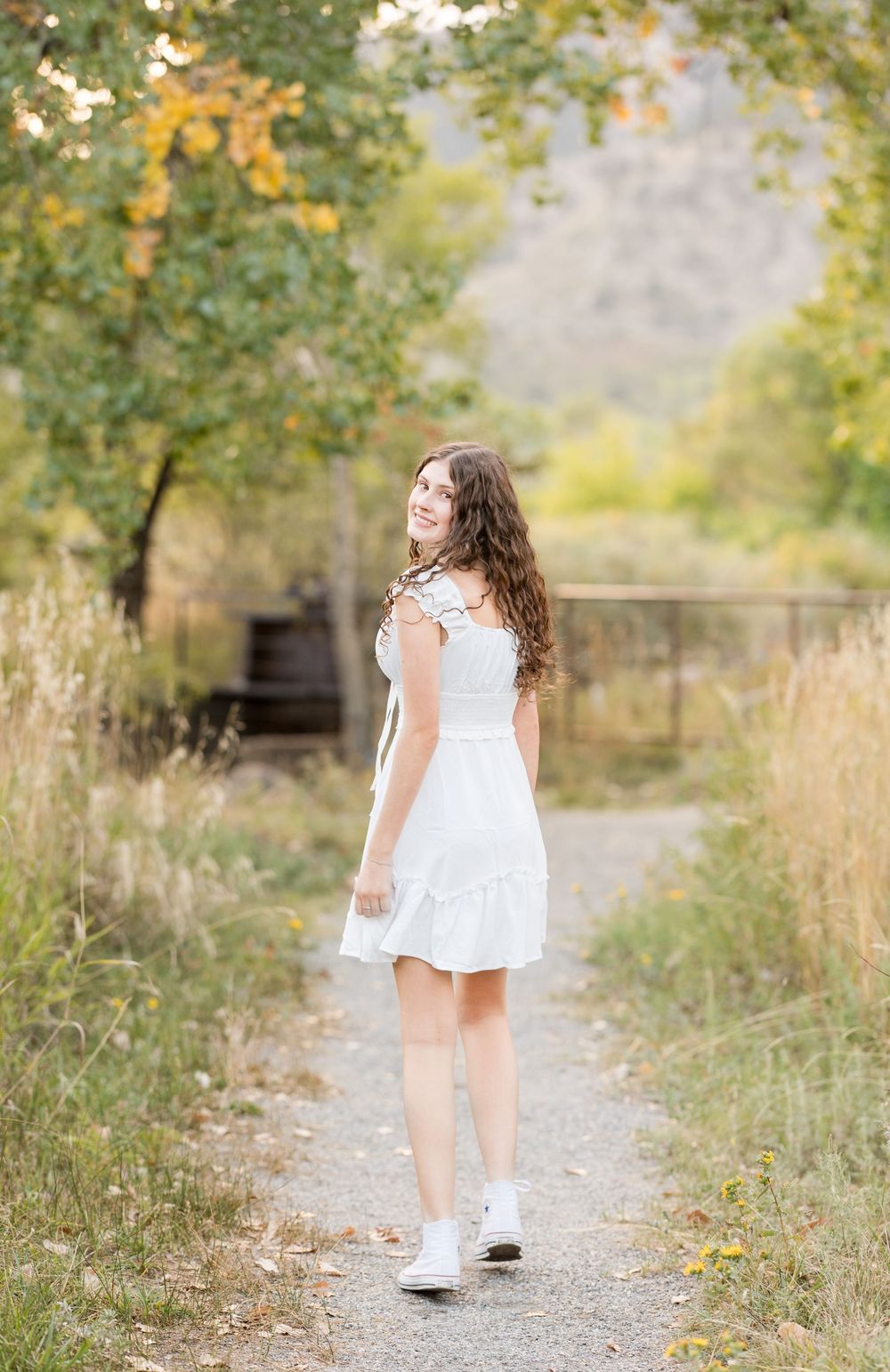 Child in white dress walking along a nature path surrounded by autumn foliage.