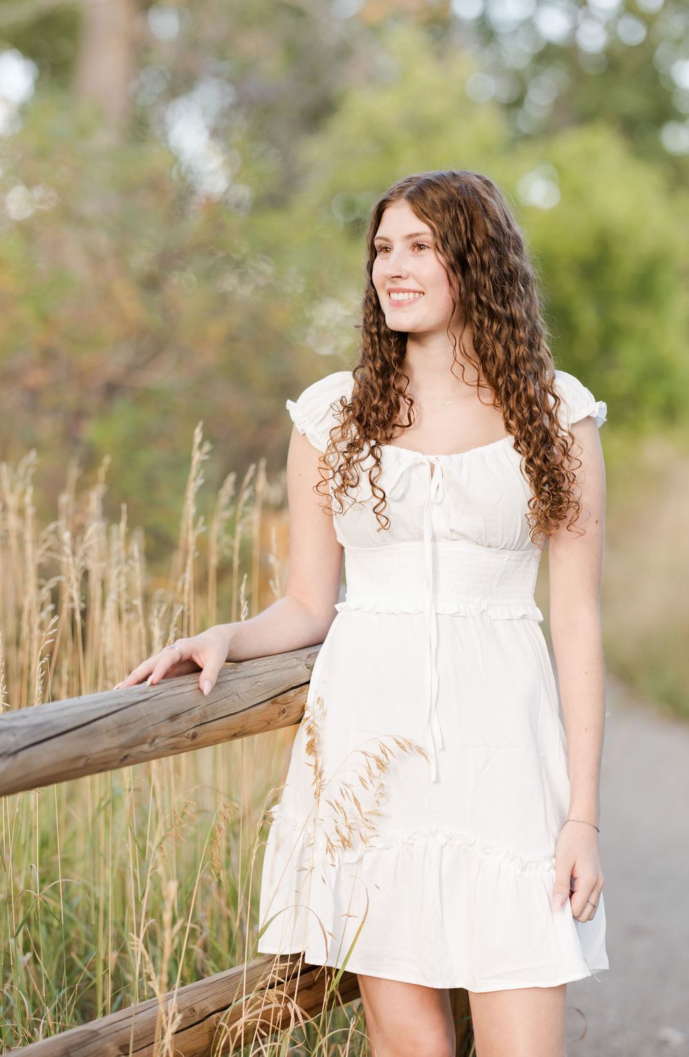 Woman in white dress poses along wooden railing with tall grass and natural outdoor setting in background.