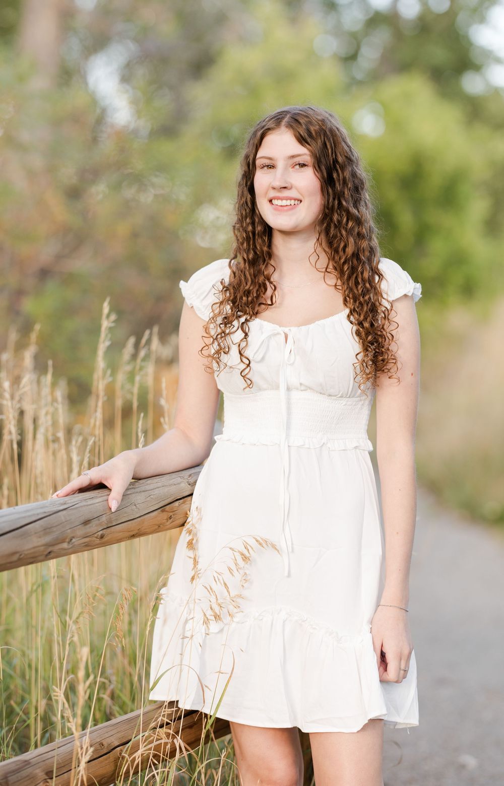 White summer dress portrait session along wooden fence with tall grass and trees in background.