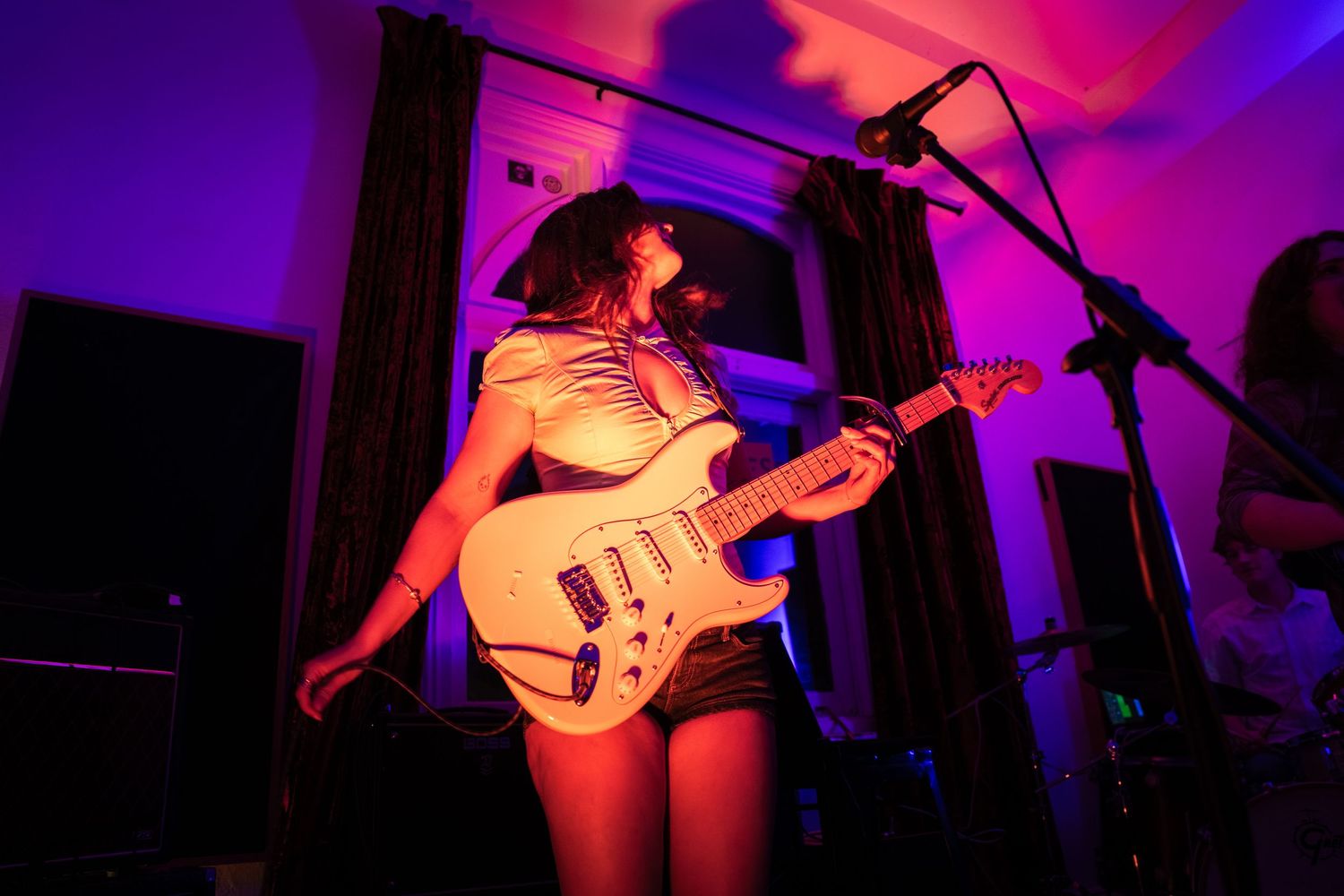 Female guitarist playing white electric guitar on stage under vibrant purple and pink stage lighting.