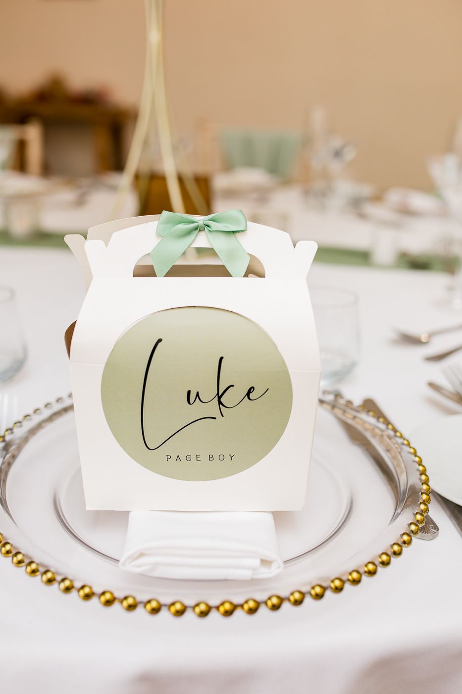 Elegant place cards with sage green accents and gold beaded charger plates on a white tablecloth.