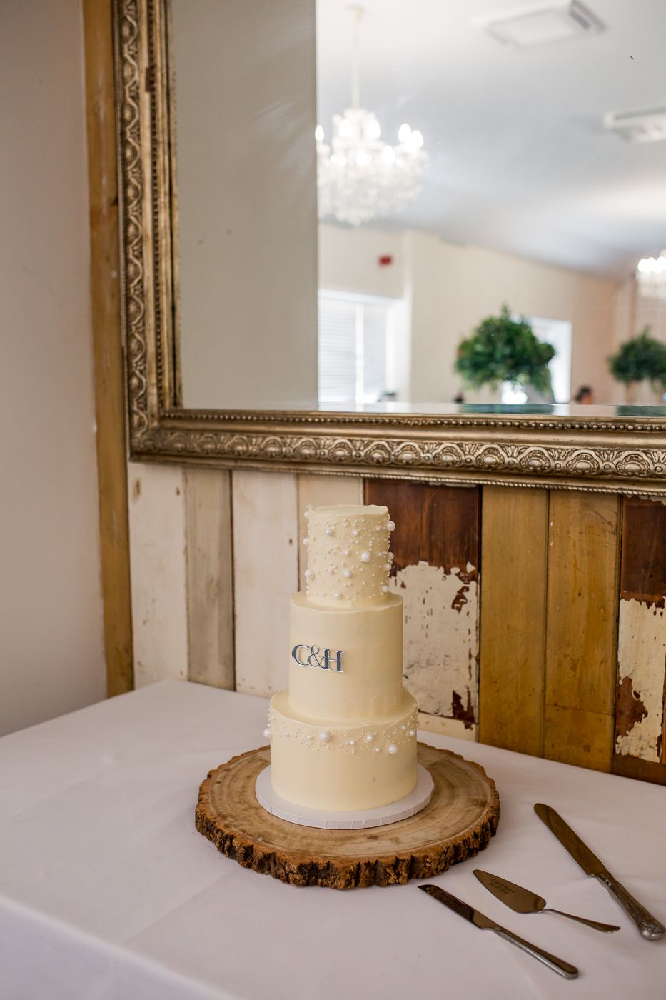 Wedding cake display setup with rustic wood slice stand and serving utensils on white tablecloth.