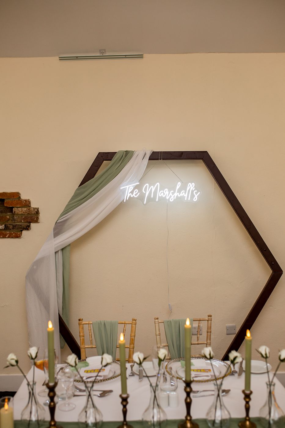 Wedding reception decor featuring white draping and neon sign with table settings in sage green and white.