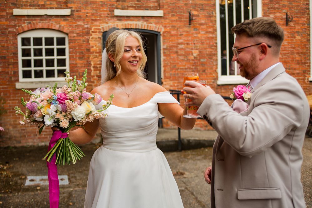 Wedding couple poses against rustic brick wall backdrop with pink floral bouquet.