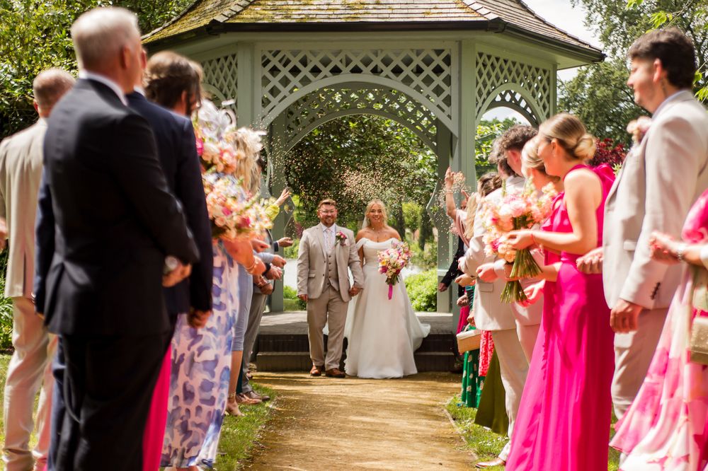 Wedding party and guests gather under garden gazebo for outdoor celebration.