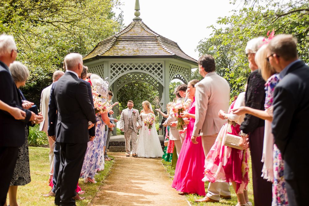 Garden wedding ceremony with guests in colorful attire under white gazebo.