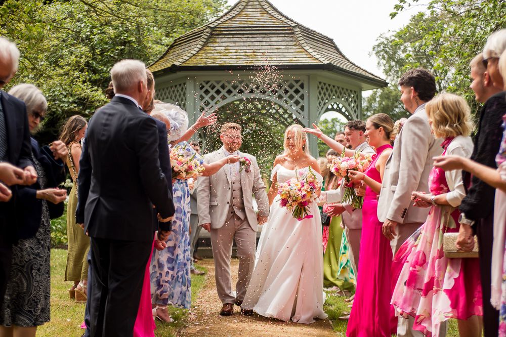 Wedding guests mingle during outdoor garden reception near white gazebo surrounded by greenery.
