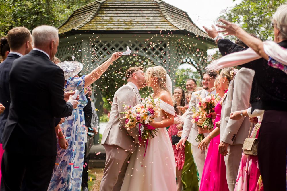 Wedding guests throw confetti during an outdoor celebration near a white gazebo.