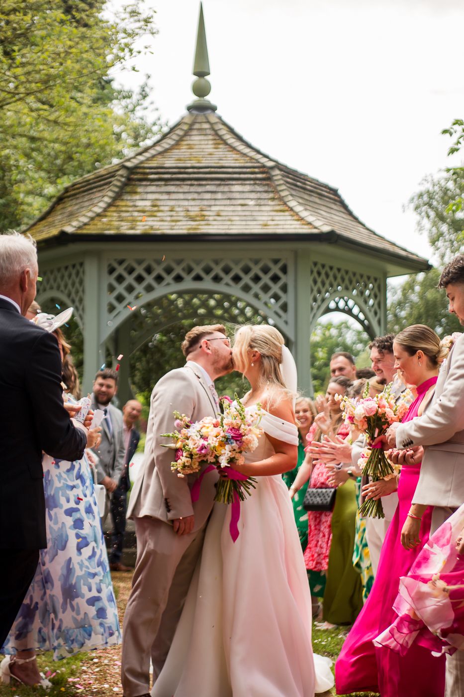 Festive wedding party gathers around a decorative garden gazebo with pink floral bouquets.