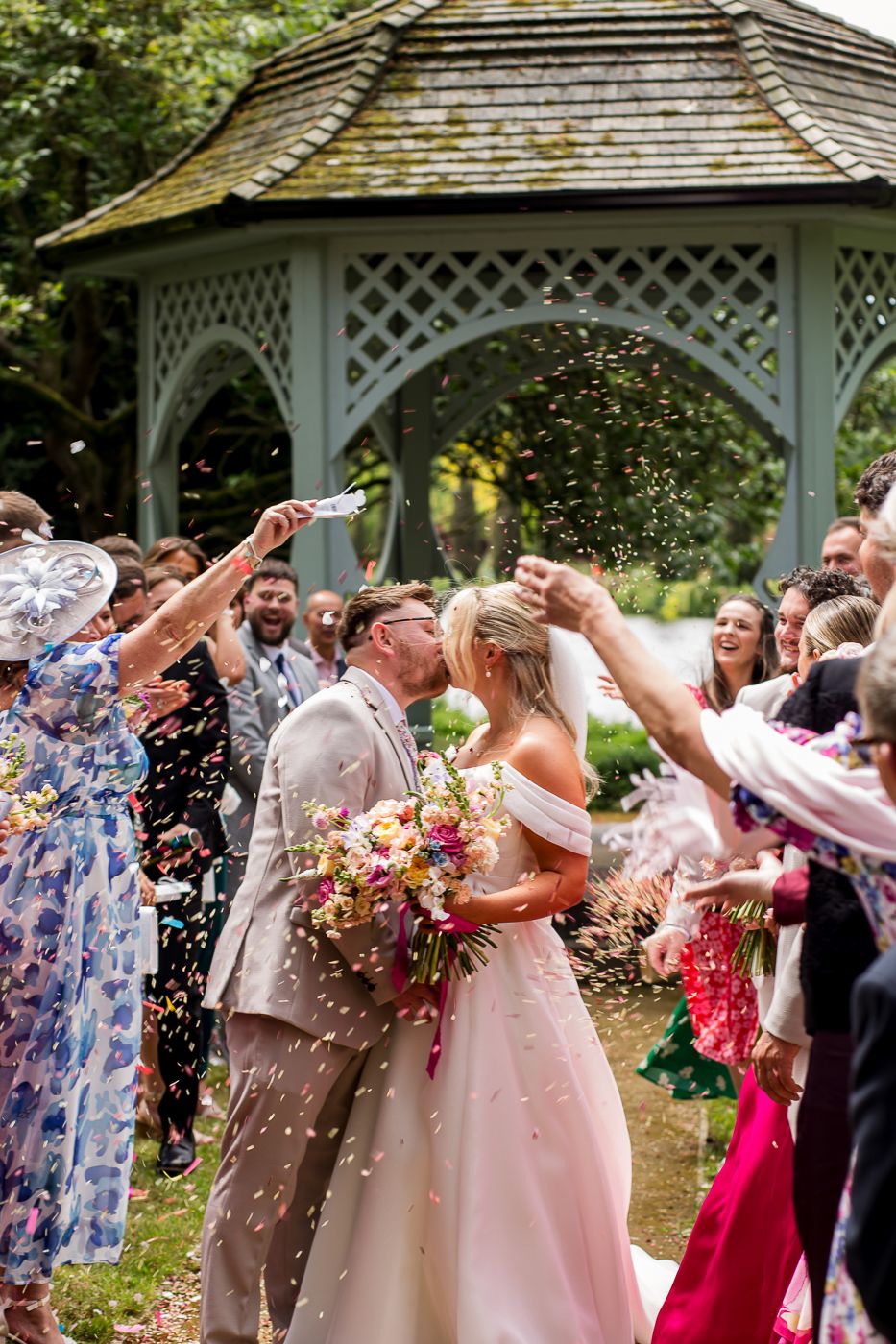 Confetti flies through the air as wedding guests celebrate near an ornate garden gazebo.