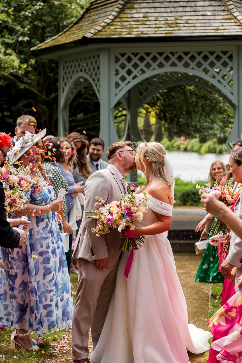 Wedding party in pink and beige attire celebrates near a white garden gazebo.