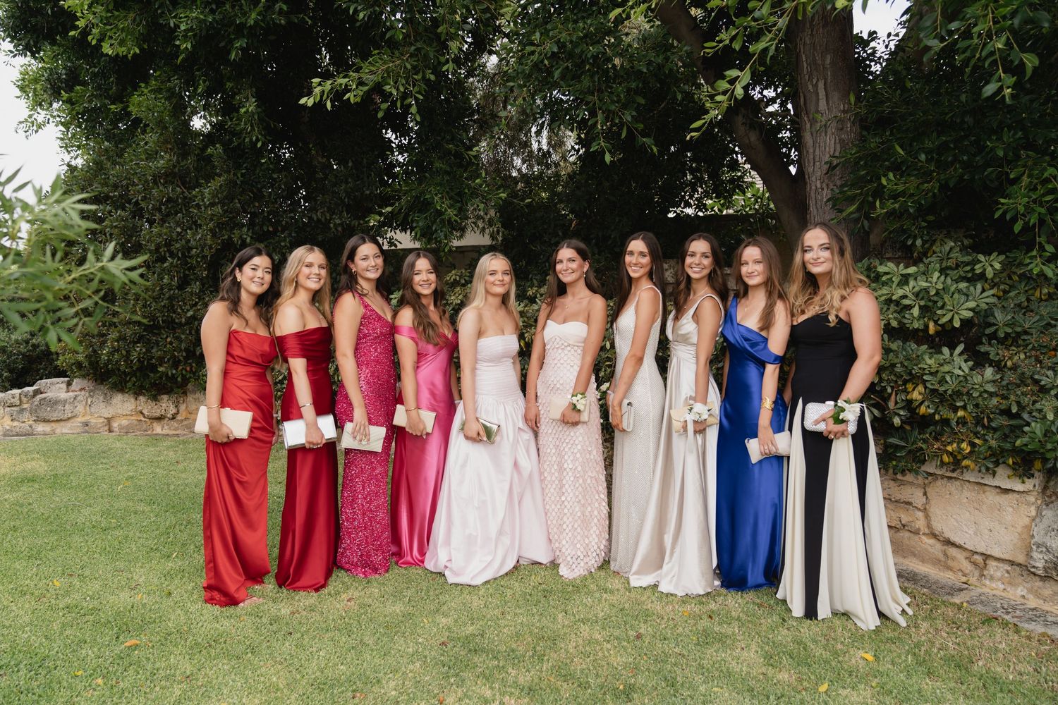 A group wearing coordinated red, pink, blue and white dresses pose together outdoors in a garden setting.