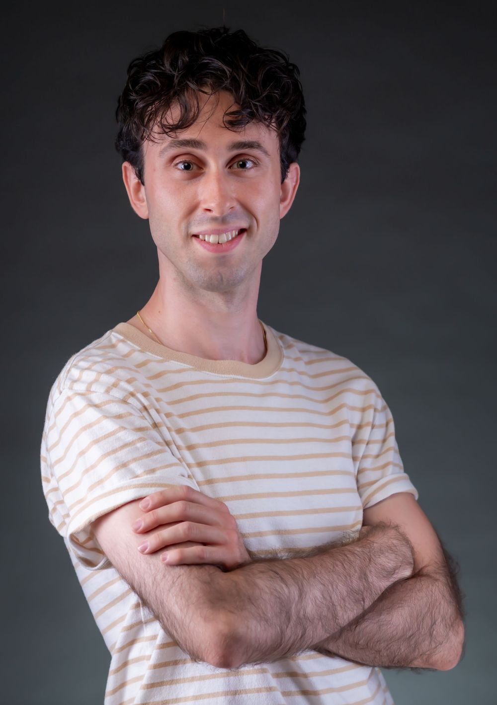 A smiling person in a striped t-shirt stands with arms crossed against a dark background.