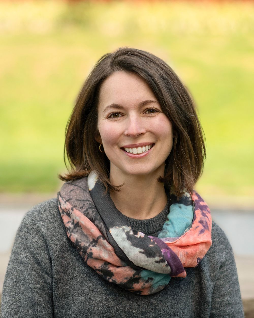 A person wearing a gray sweater and colorful floral scarf smiles warmly in an outdoor setting.