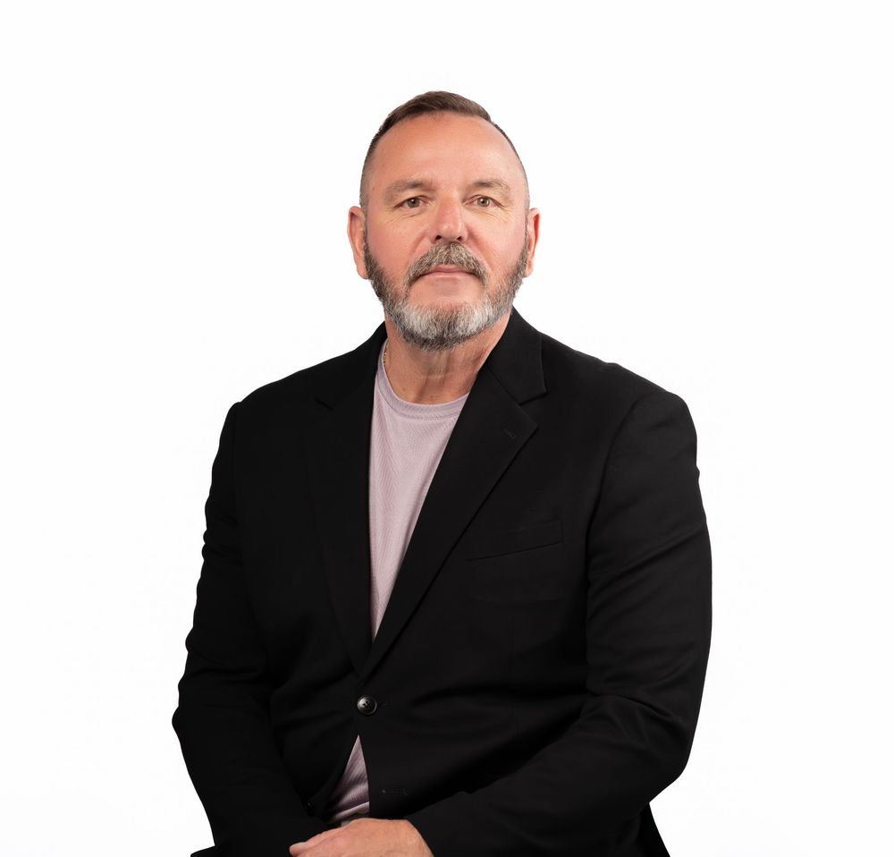 Professional headshot of someone in a black suit and pink dress shirt against white background.