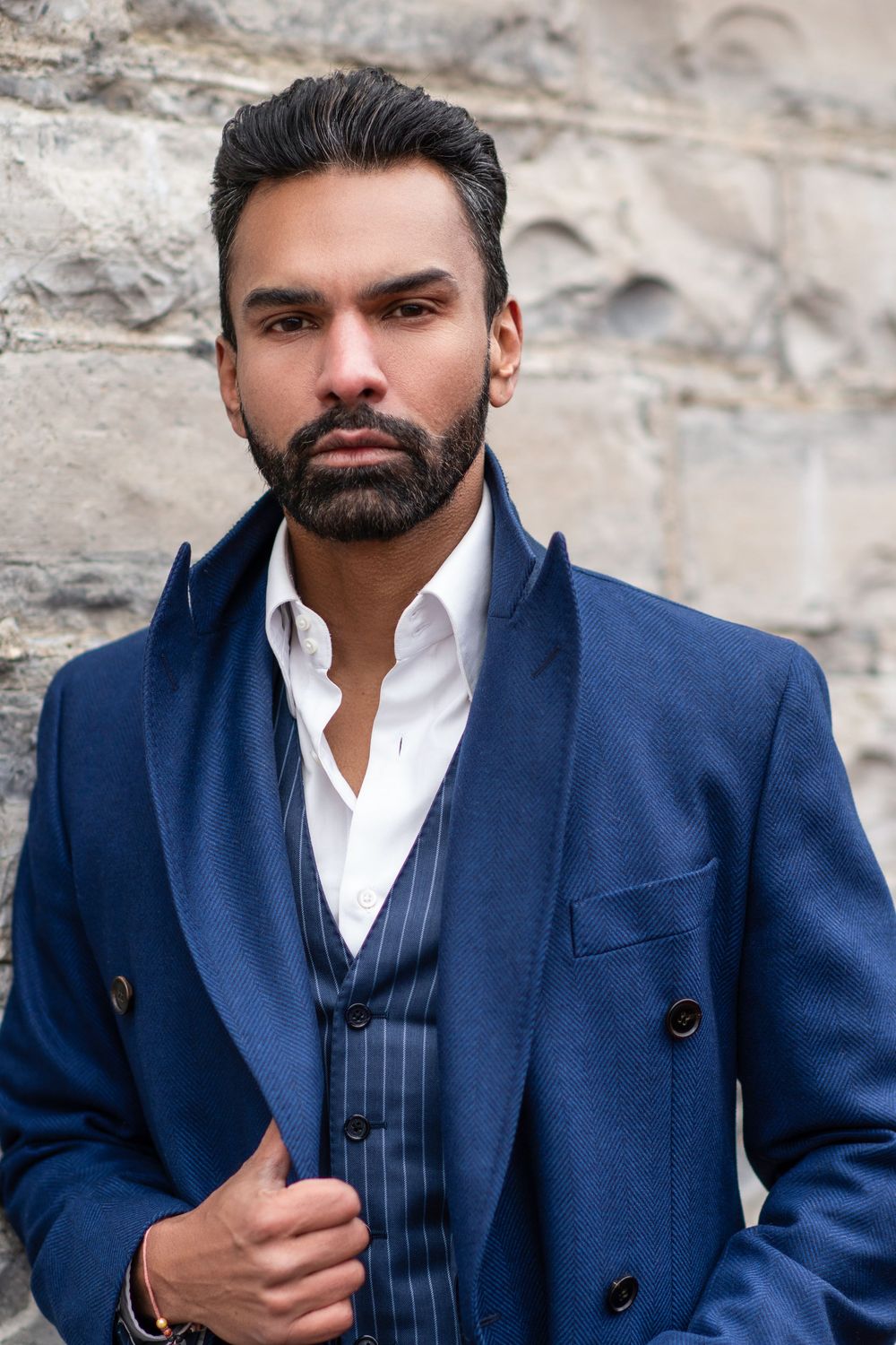 A person in a stylish navy blue suit and white dress shirt poses against a textured stone wall.
