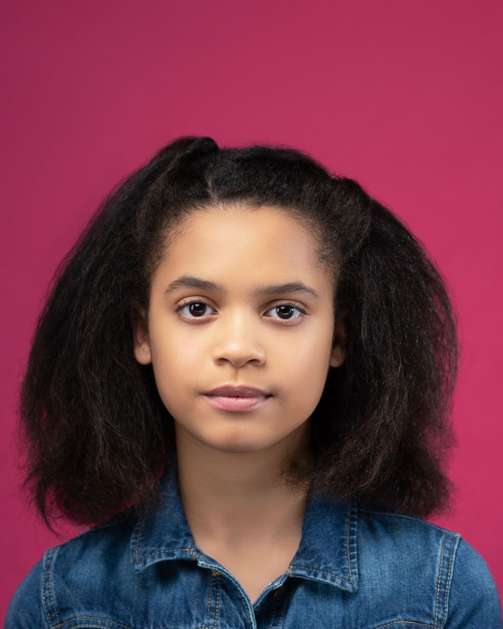 Person with natural curly hair in denim jacket poses against vibrant pink background.