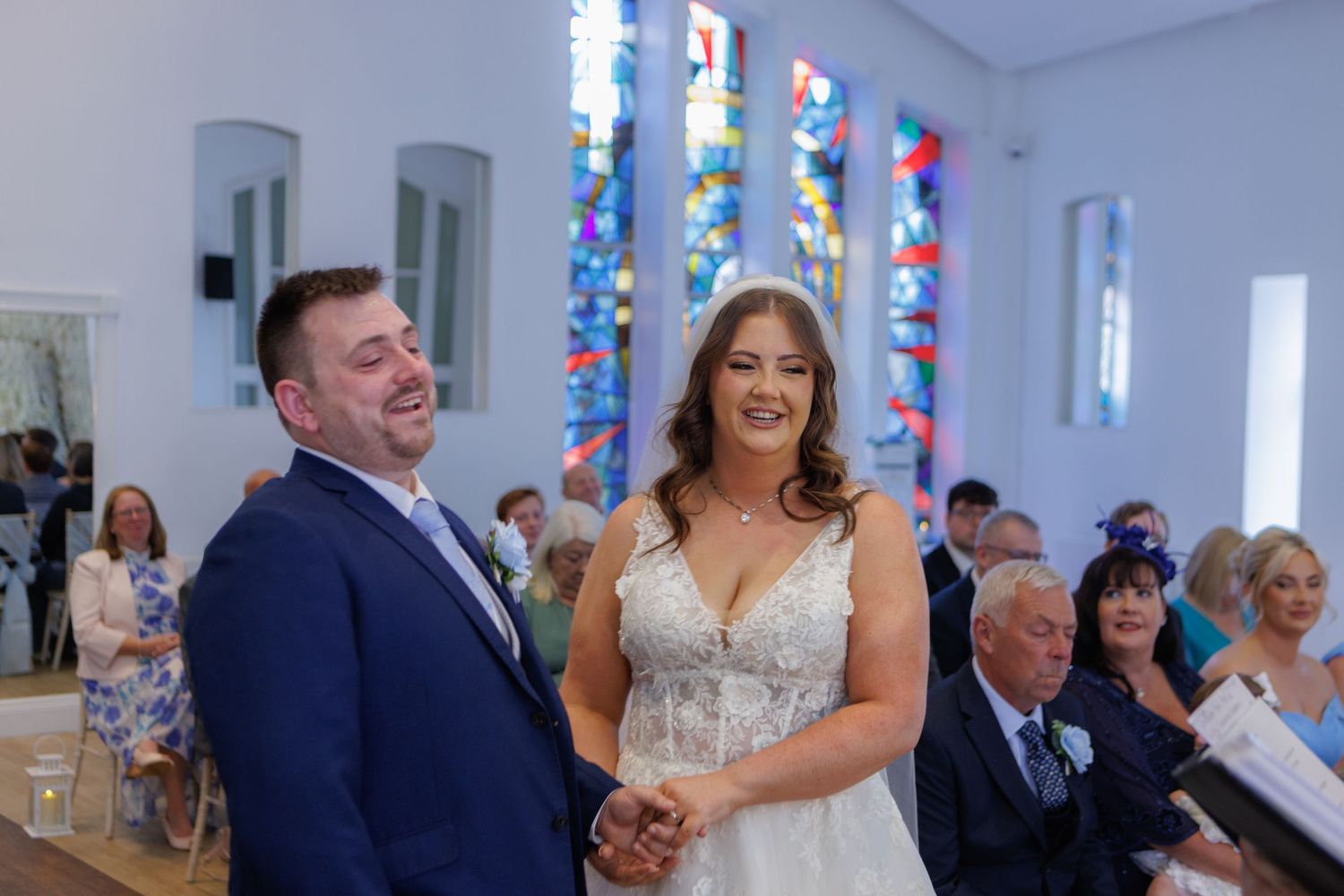 Couple shares a joyful laugh during wedding ceremony inside chapel with colorful stained glass windows.