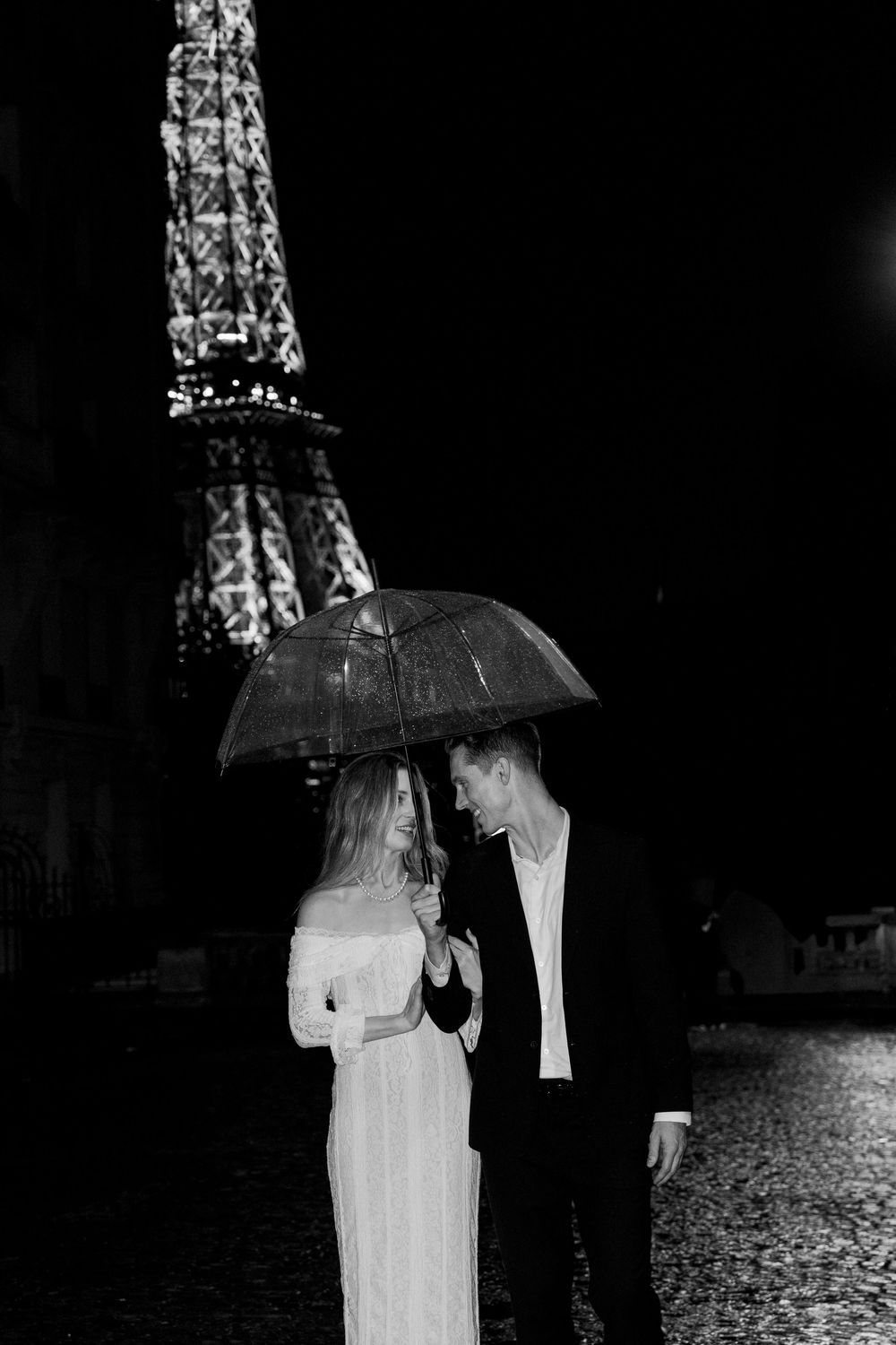 A couple shares an umbrella in the rain near the illuminated Eiffel Tower in Paris at night. Paris film photographer.
