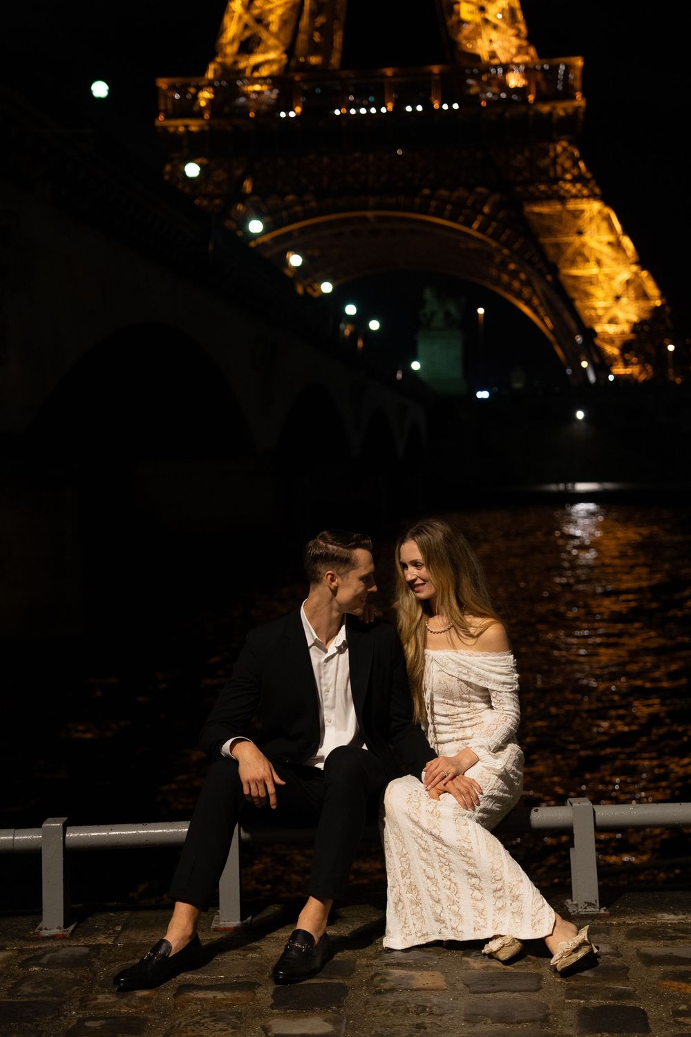 A romantic couple sits along the Seine River at night with the illuminated Eiffel Tower glowing in the background. Paris editorial elopement