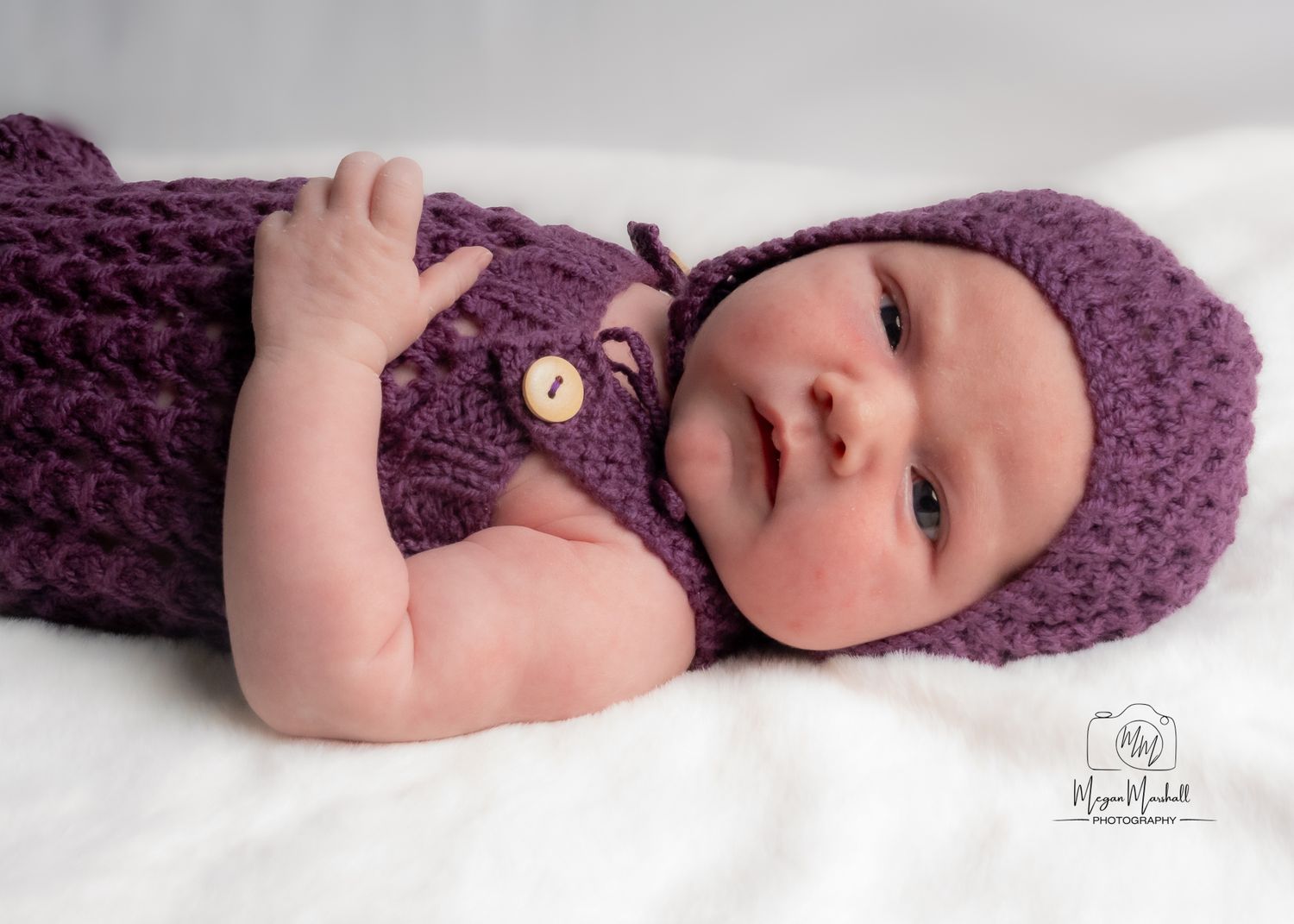 Newborn Photography - Newborn baby wearing purple knitted hat and sweater lying on white blanket.