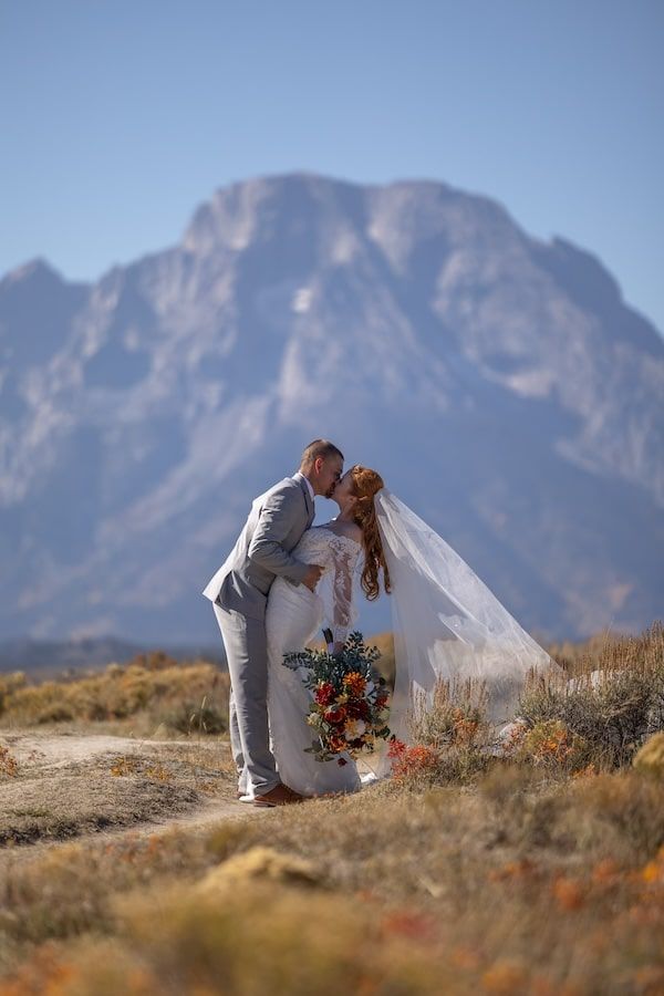 A couple in wedding attire embraces in front of majestic mountain scenery in a desert landscape.