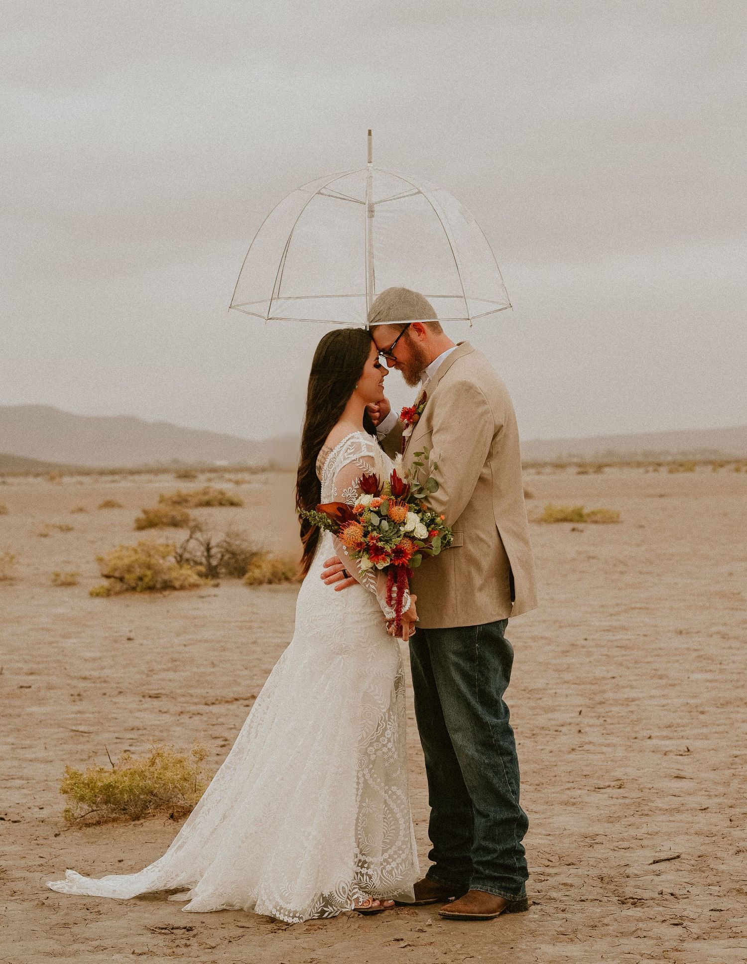 A couple shares an intimate moment in the desert during their wedding photoshoot, with a soft vintage-toned backdrop.
