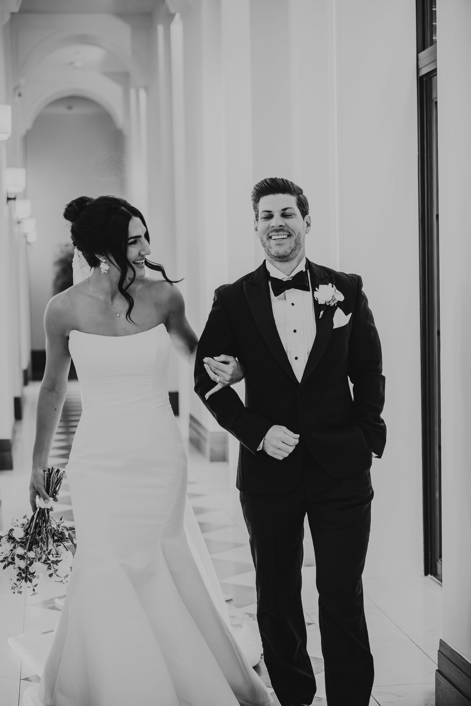 Black and white wedding photo of bride in strapless gown and groom in tuxedo sharing a candid moment in hallway.