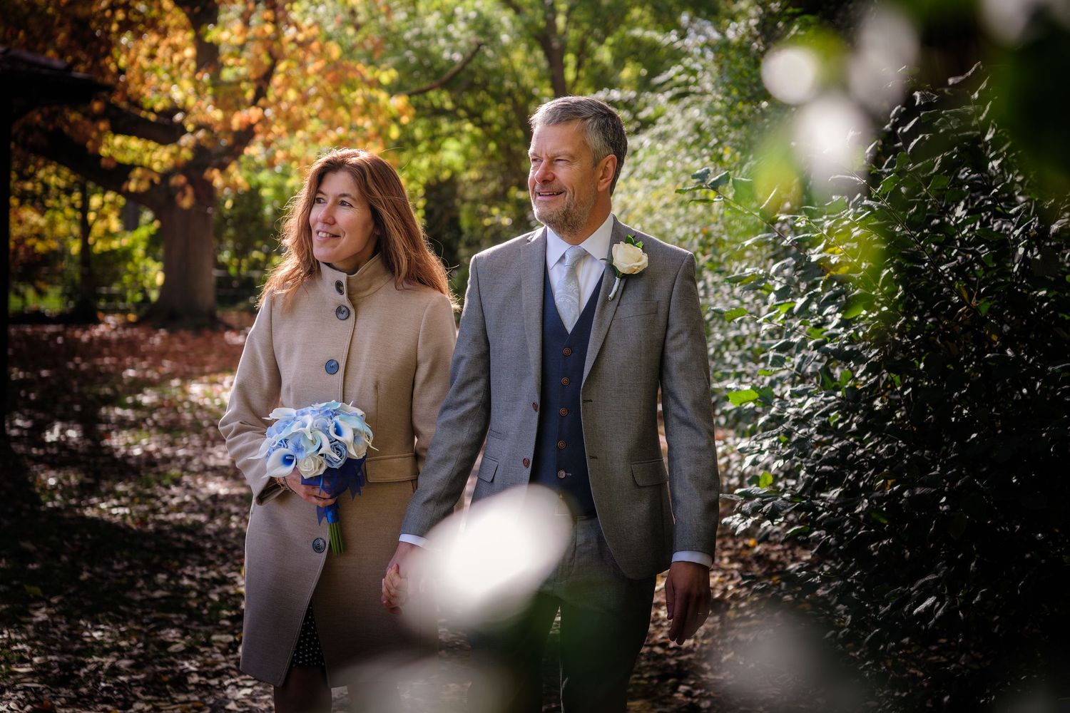 A wedding couple walks together through a lush garden setting with sunlight filtering through the trees.