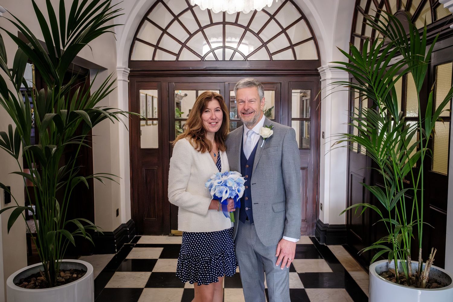 A couple stands in front of a black and white checkered floor with tropical plants and an arched doorway.