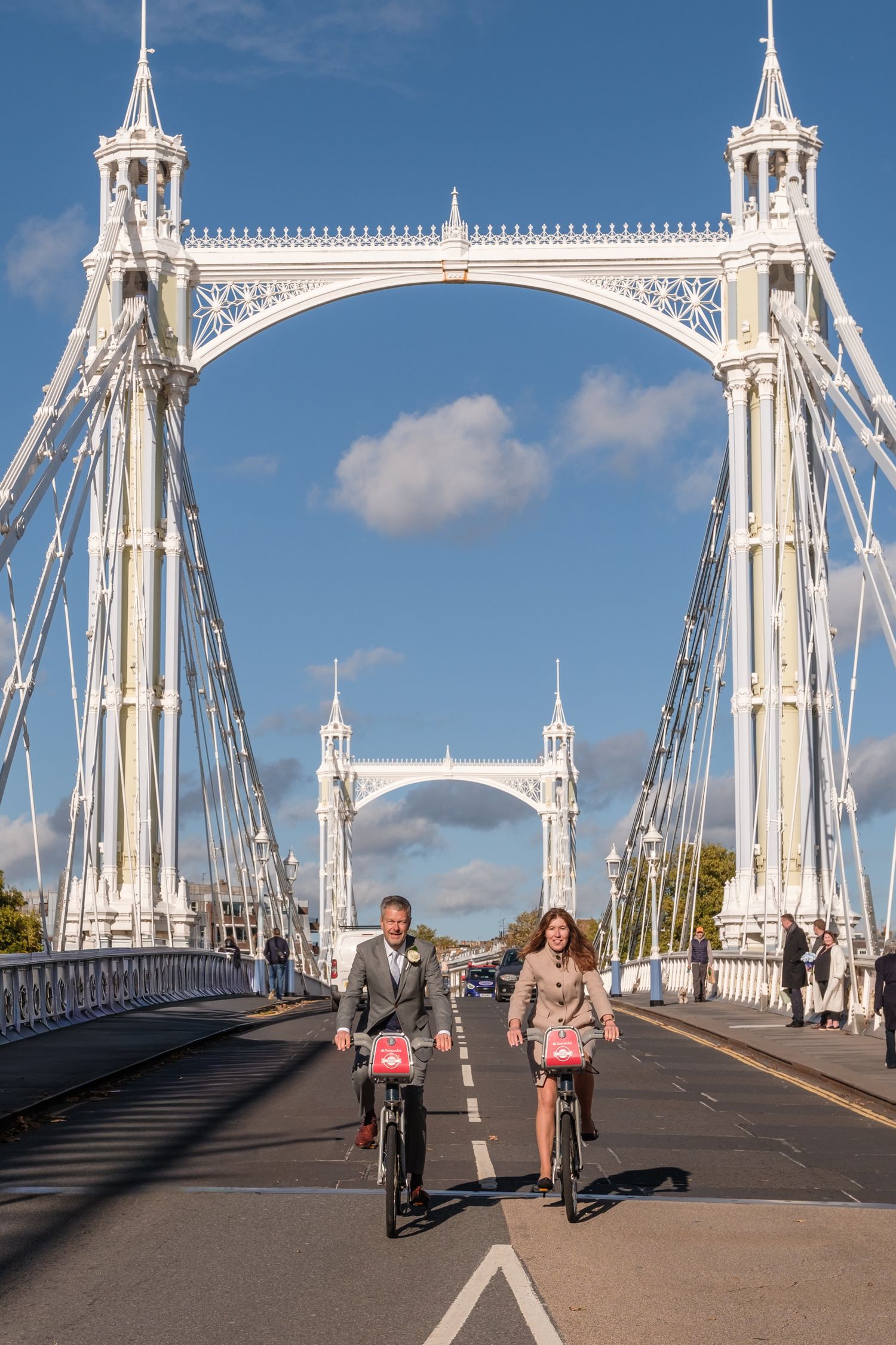 People walking and cycling across the iconic Albert Bridge in London on a sunny day with white ornate architecture.