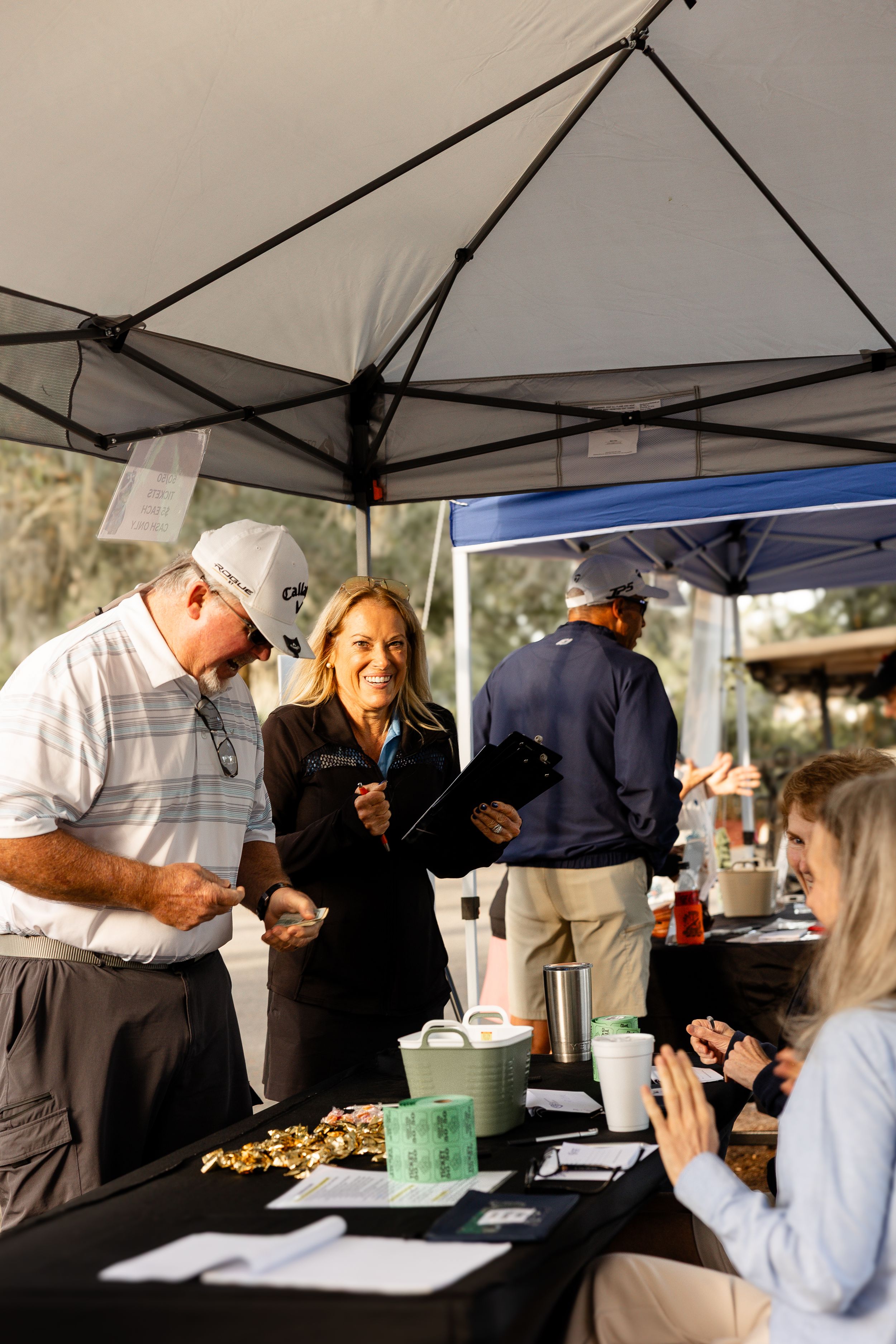People gathering under white canopy tents at an outdoor event with food and refreshments.