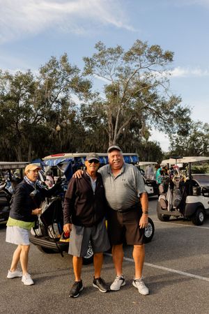 Golfers gathered in a parking lot with golf carts and trees in the background on a sunny day.