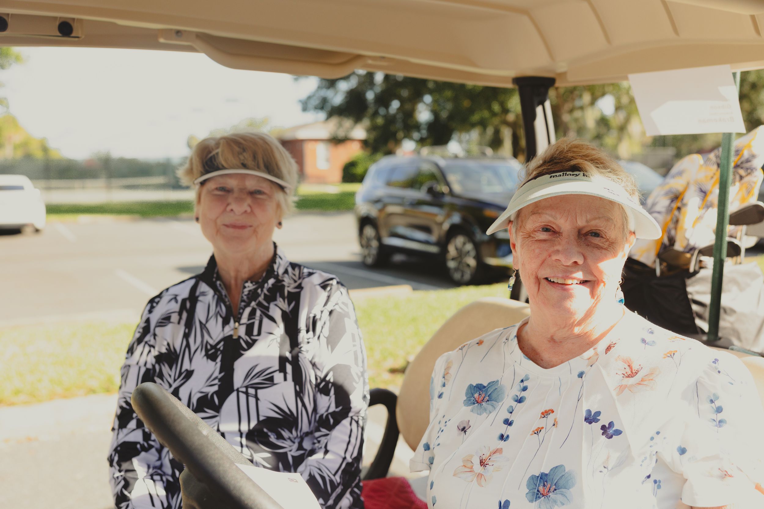Two people sit together in a golf cart wearing patterned shirts on a sunny day.