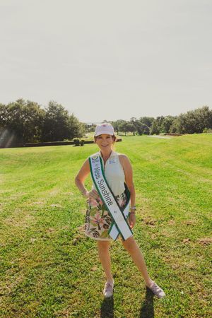 A golfer wearing a sash poses on a sunny golf course with trees in the background.