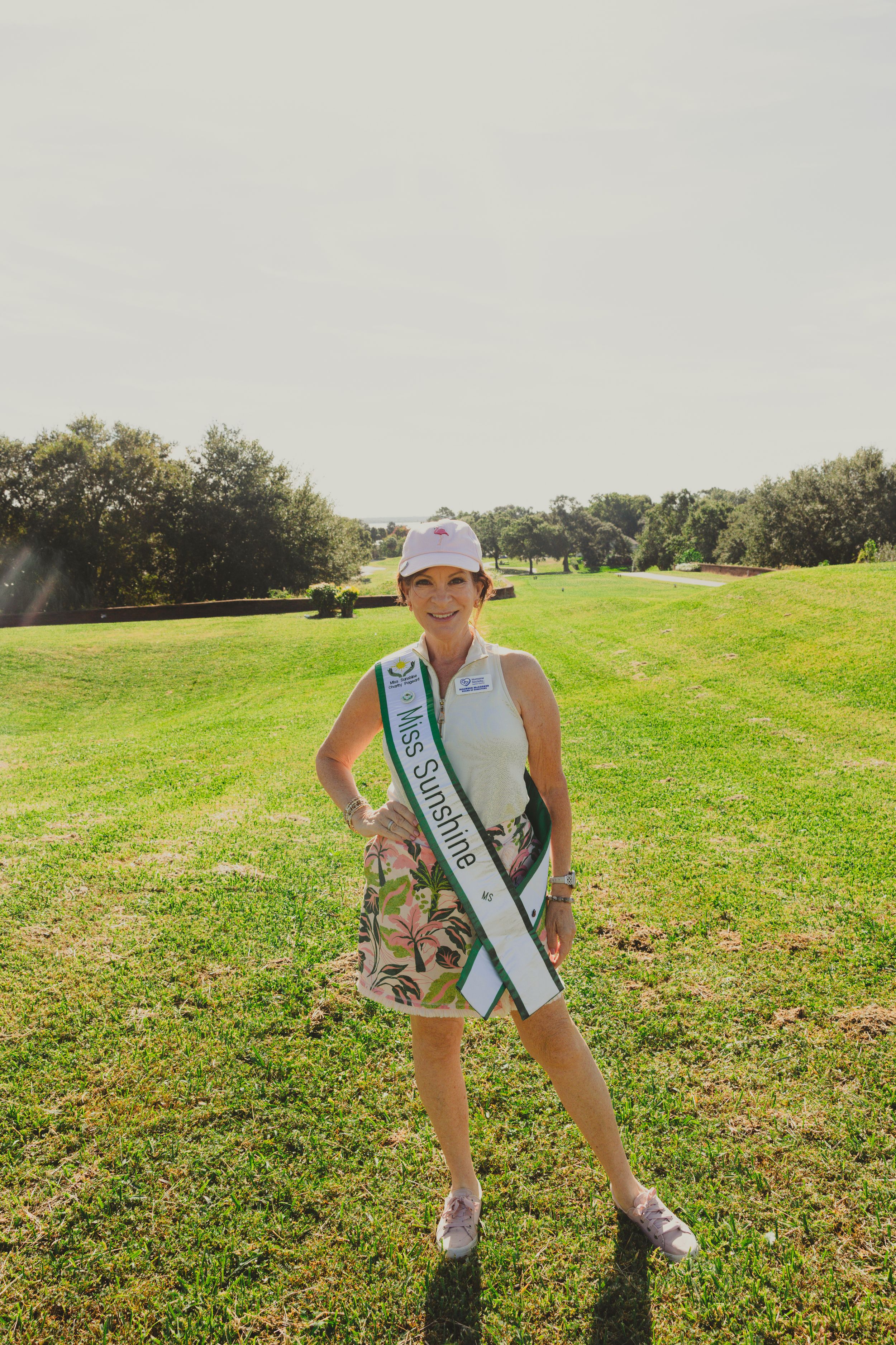 A golfer wearing a sash poses on a sunny golf course with trees in the background.