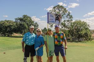 Group of golfers gathered on a lush green course under blue skies with palm trees in the background.