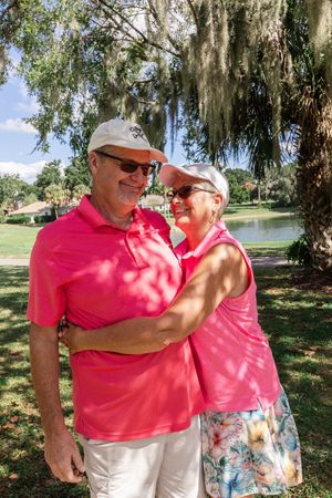 Two people wearing matching pink shirts embrace while standing outdoors on a sunny day near trees.