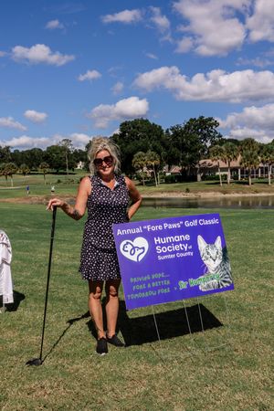A person in a polka dot dress poses with a purple Humane Society banner on a sunny golf course.