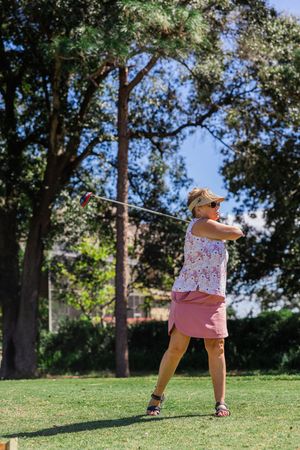Person in pink shirt and shorts practicing golf stance on grassy hill under blue sky.