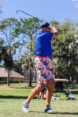 Golfer in blue shirt and colorful patterned shorts demonstrating golf swing technique on grassy course.