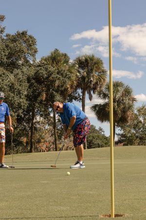 Golfers practicing their putting skills on a sunny golf course with palm trees and yellow flag markers.