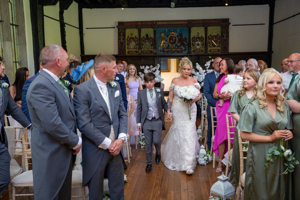 Bride walks down the aisle escorted by family in an elegant wood-paneled wedding venue.
